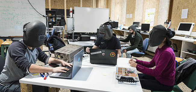 Des étudiants avec un casque de protection travaillent sur des ordinateurs.